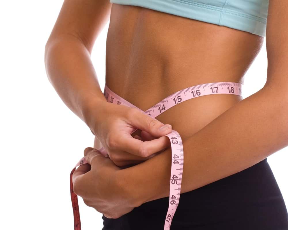 Person in exercise wear measuring their waist with a pink measuring tape, wearing a light blue crop top and black bottoms.