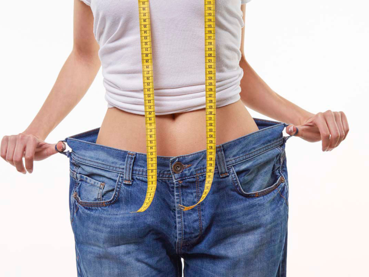 Alt text: Person in a white shirt pulling out loose-fitting jeans with a measuring tape hanging down, demonstrating weight loss.